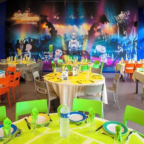 Um restaurante com mesas redondas, cadeiras verdes e laranja, e um cenário colorido com tema de robôs.