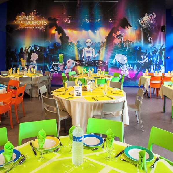A restaurant with round tables, green and orange chairs, and a colorful robot-themed backdrop. - Hotel du Futuroscope (Chasseneuil-du-Poitou)