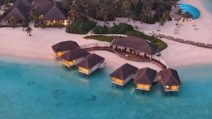 An aerial view of overwater bungalows with thatched roofs, connected by a walkway, on a beach with clear turquoise water. - Nova Maldives (Malé)