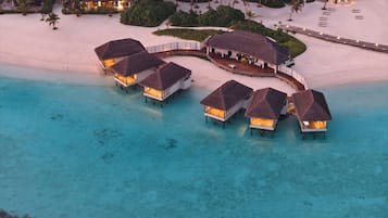An aerial view of overwater bungalows with thatched roofs, connected by a walkway, on a beach with clear turquoise water.