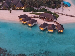 Vista aérea de bungalows sobre el agua con techos de paja, unidos por un paseo, en una playa con aguas turquesas cristalinas.