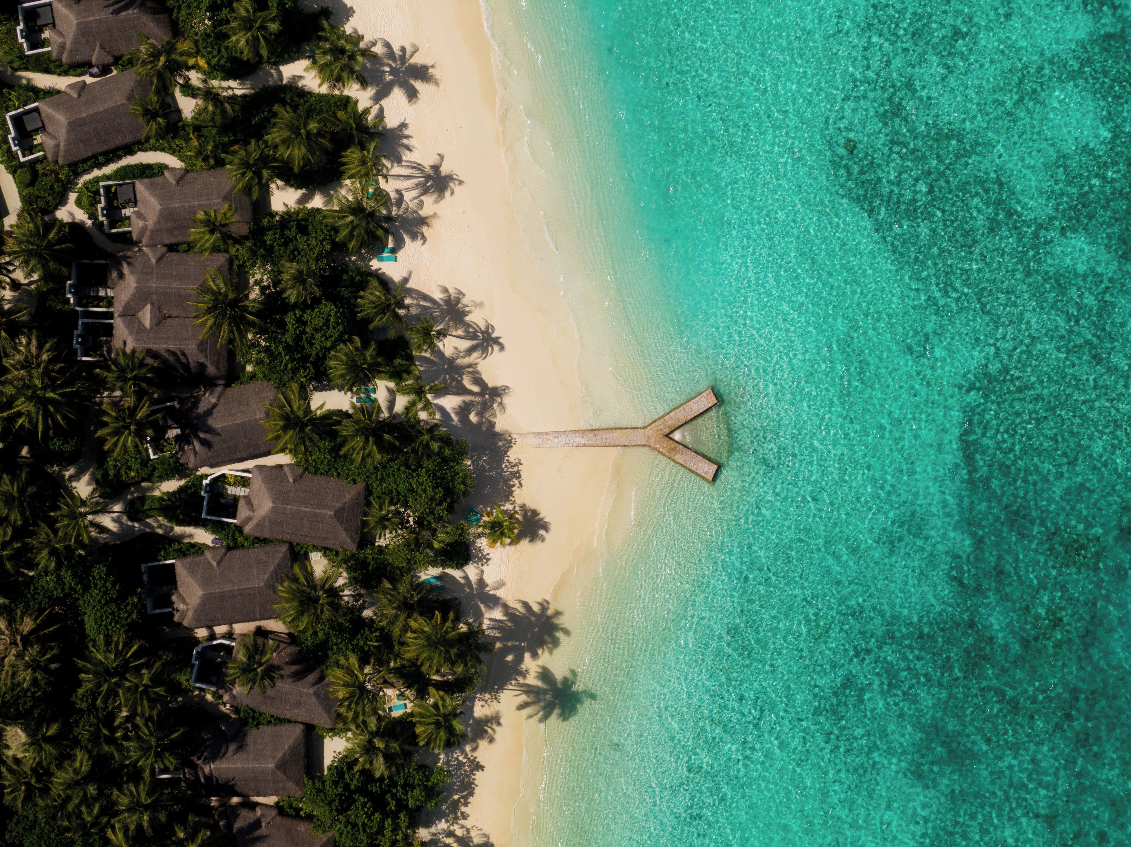 An aerial view of a beach resort with overwater bungalows, clear turquoise waters, and palm trees.