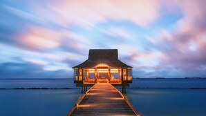 A wooden overwater structure with a long pier leading to it, surrounded by calm water and a twilight sky. - Nova Maldives (Malé)