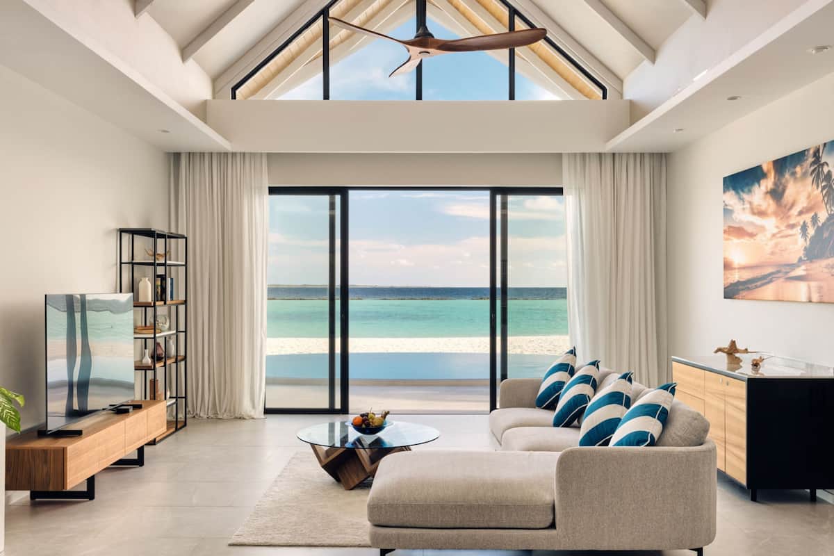 A modern living room with a large window overlooking a beach, a sofa with cushions, a coffee table, and a TV on a wooden console.