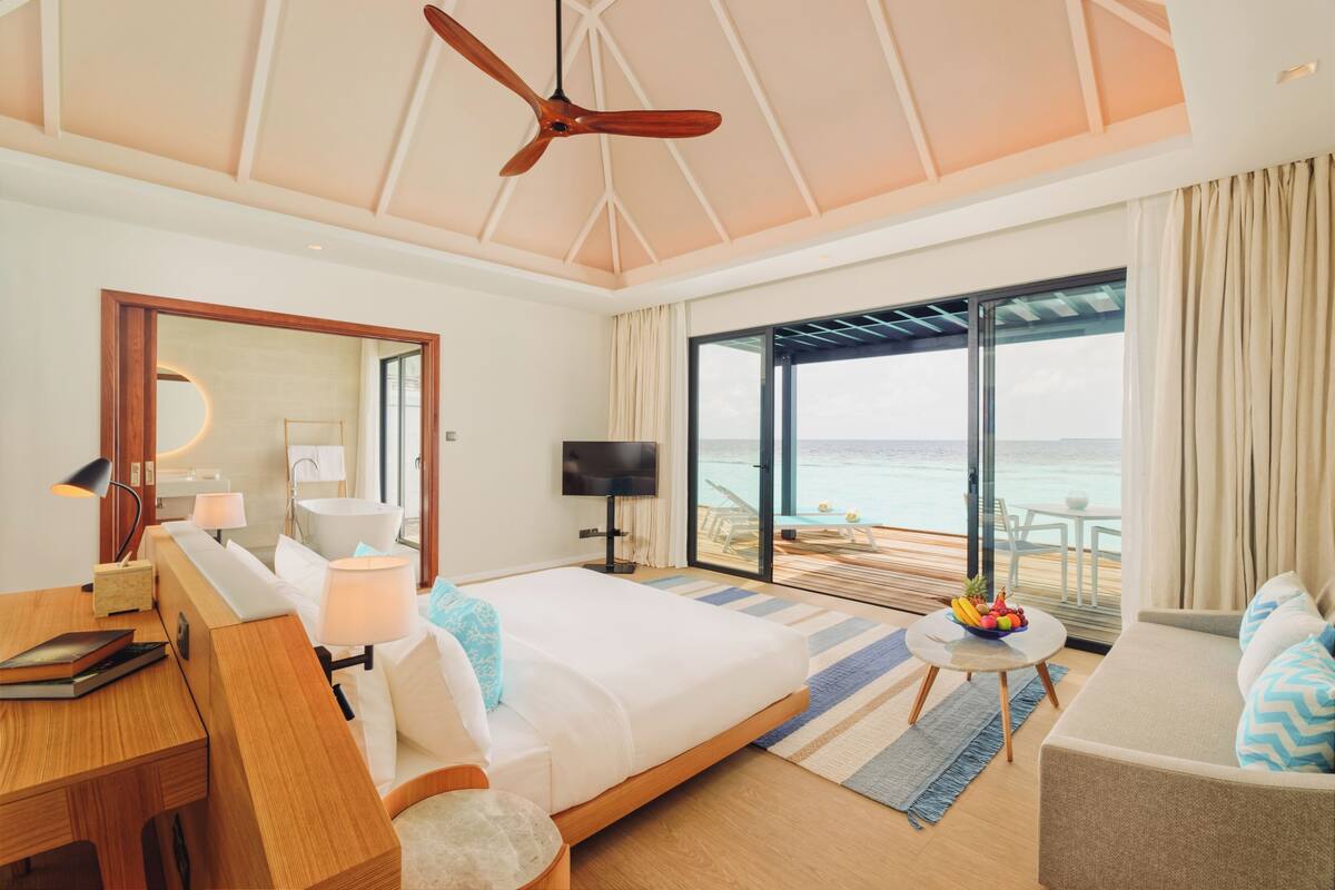 A bedroom with a bed, a desk, a TV, and a balcony with a view of the sea.