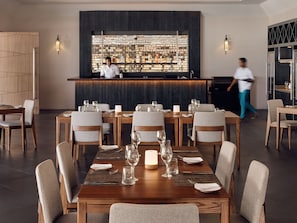 Un restaurante moderno con mesas y sillas de madera, una zona de bar, y una persona caminando al fondo.