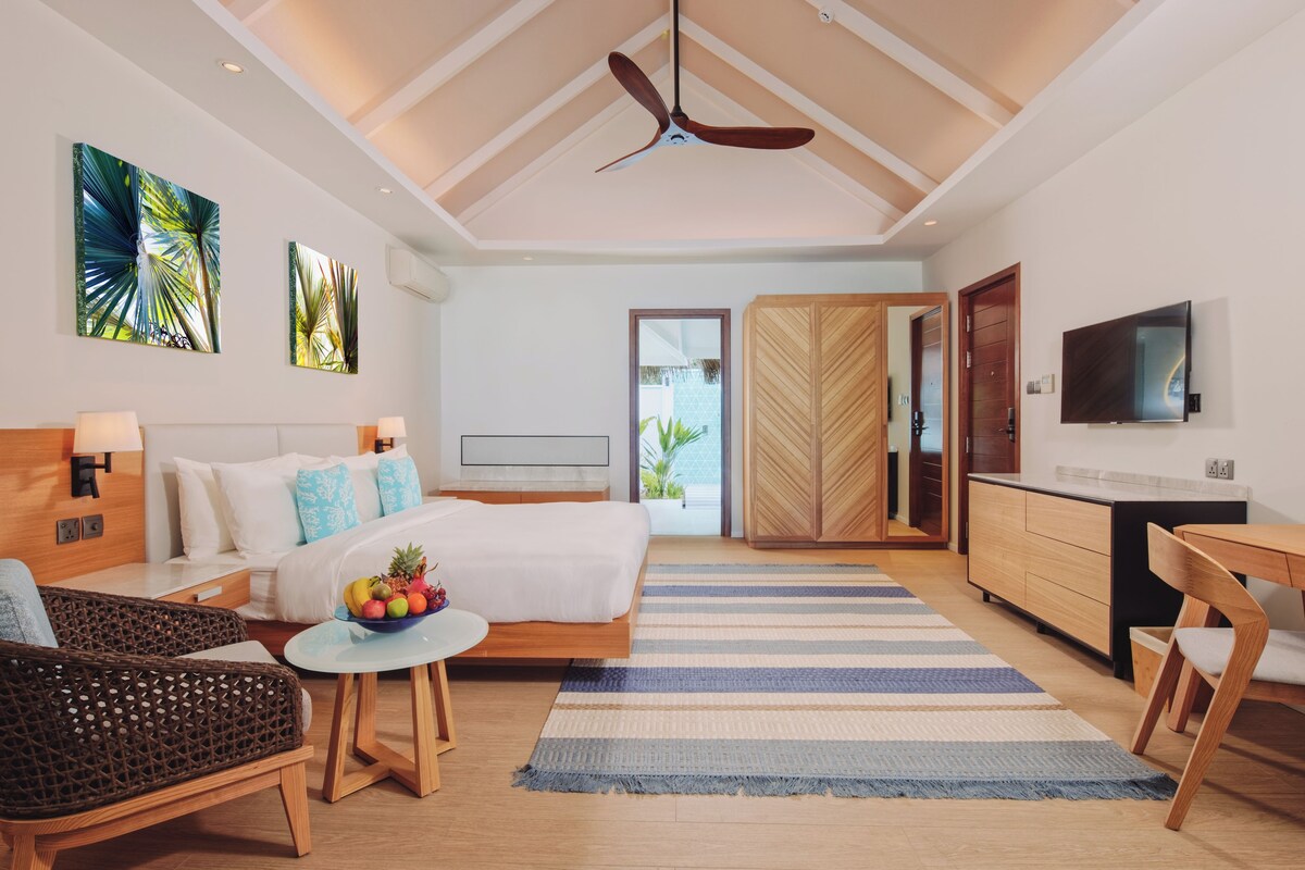 A modern hotel room with a bed, a dining area, a TV, and a view of the outdoors.