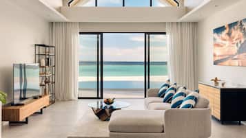 A modern living room with a large window overlooking a beach, a sofa with cushions, a coffee table, and a TV on a wooden console.