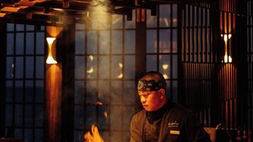 Un chef cuisine à une table haute, entouré de flammes, dans un restaurant à la décoration traditionnelle et à l’ambiance chaleureuse.