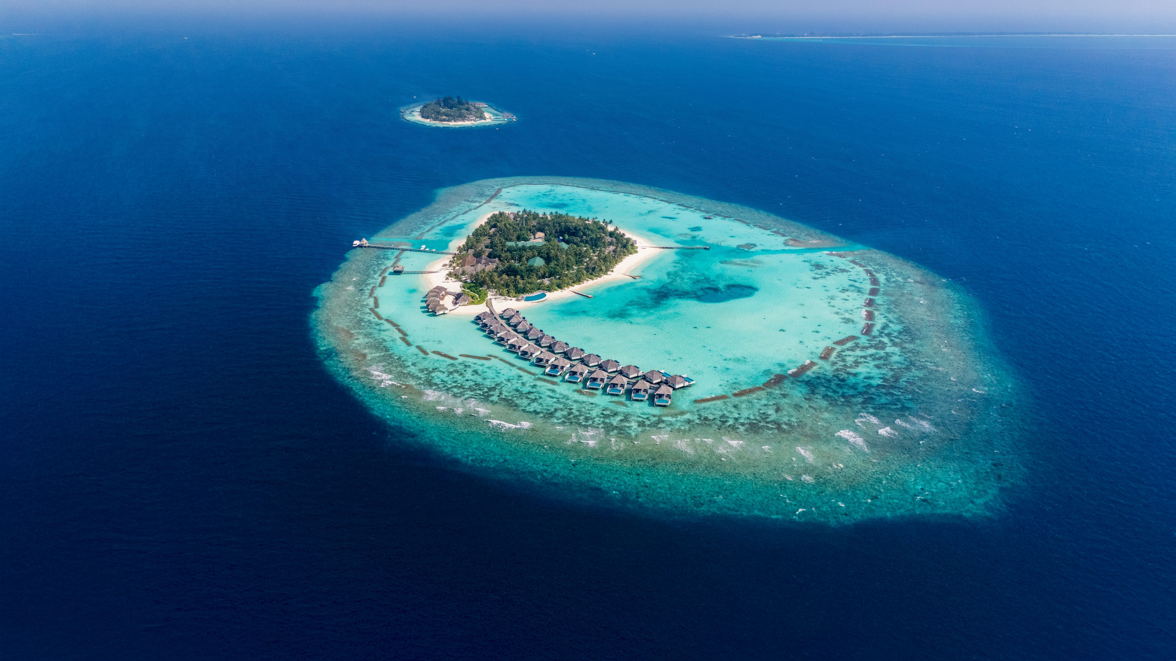 An aerial view of a tropical island with a crescent-shaped resort area featuring overwater bungalows and a central building.