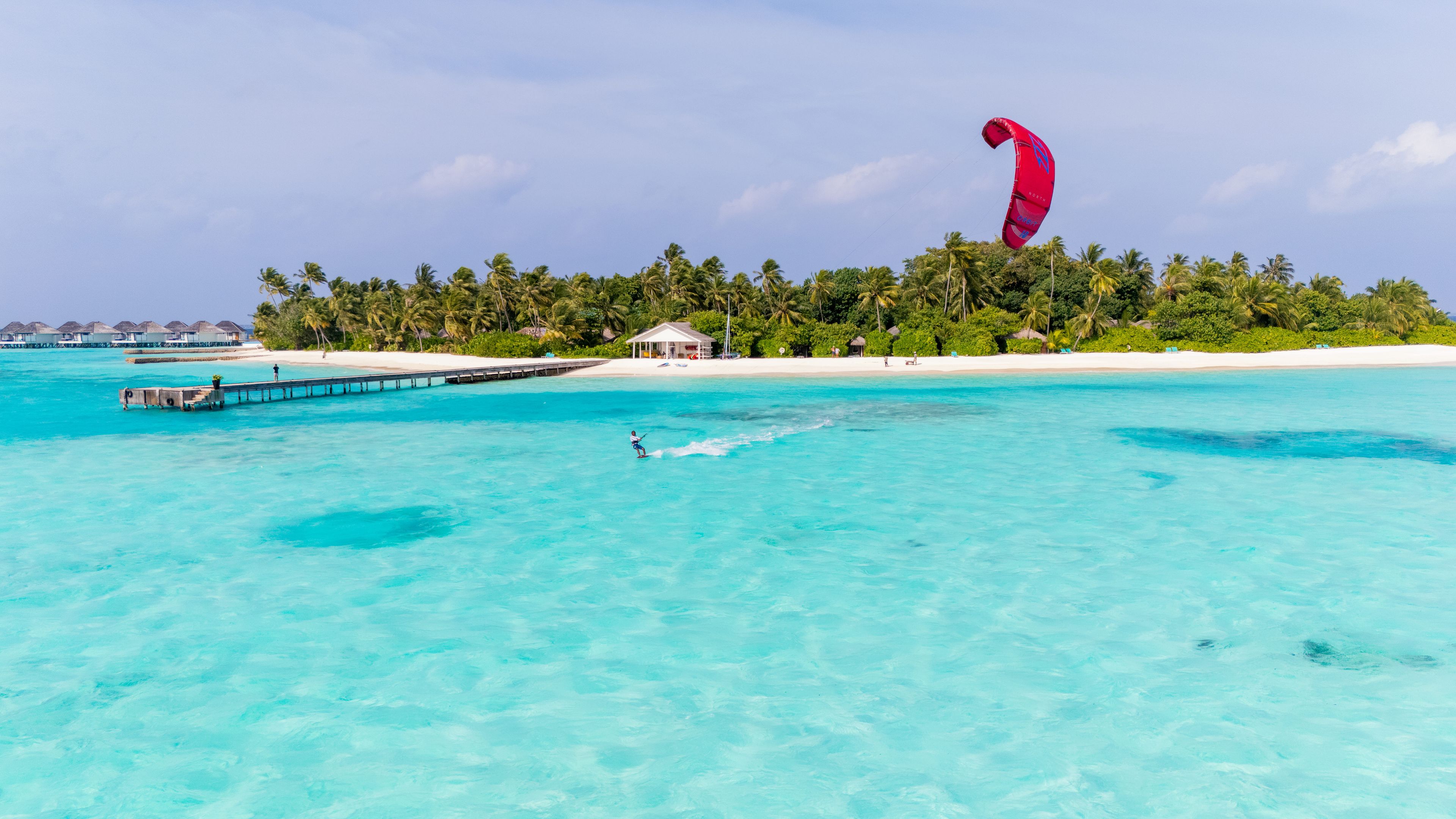 A beach with clear turquoise water, a pier, and a person kite surfing.