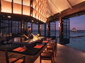 A restaurant with a chef preparing food, a bar area, and a view of the sea.