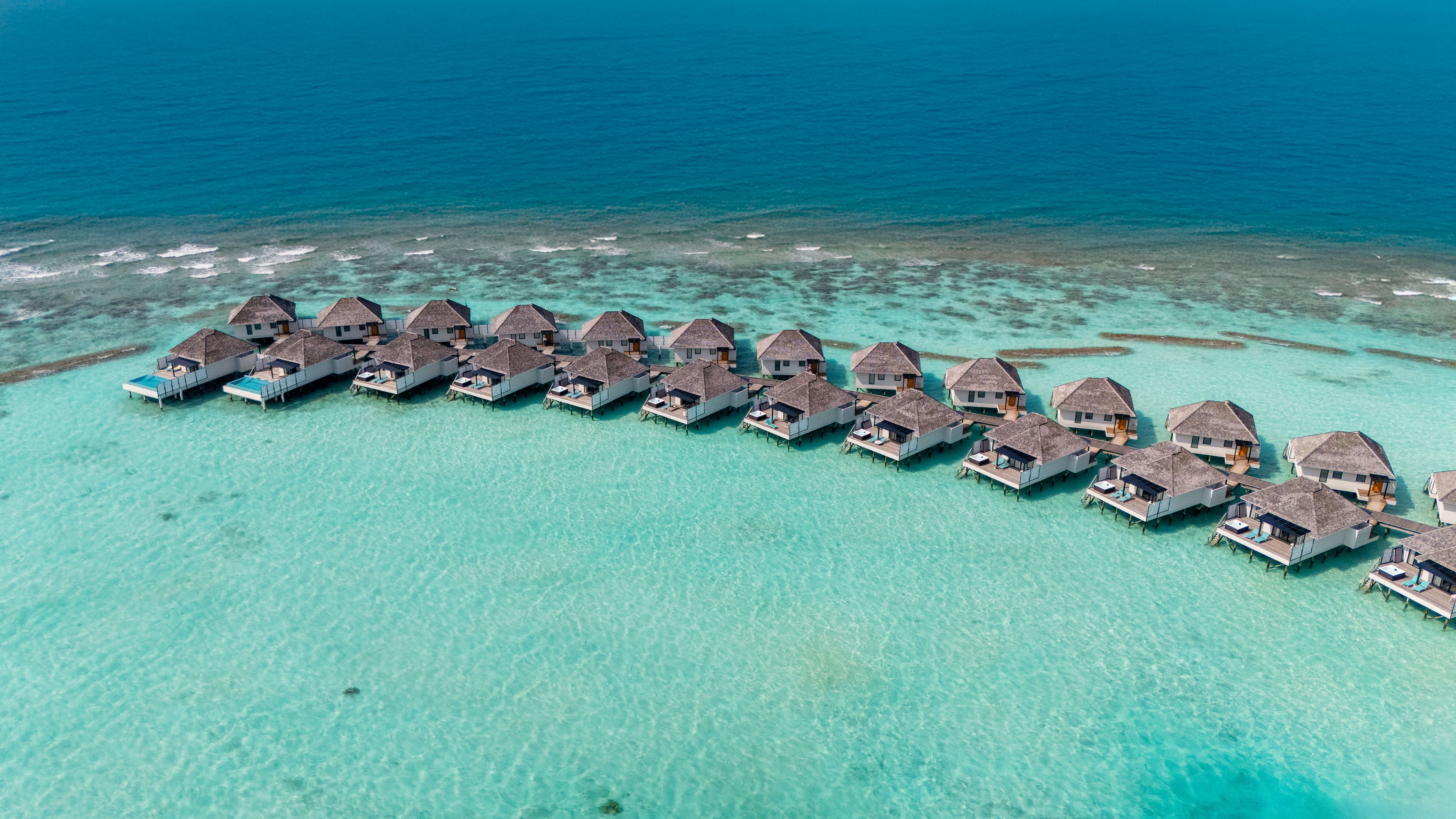An aerial view of overwater bungalows aligned in a row on a turquoise coral reef, connected by walkways.