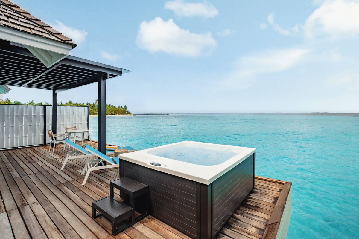 A wooden deck with a hot tub, lounge chairs, and a covered seating area overlooking a clear blue sea.