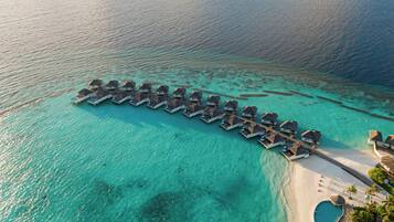 An aerial view of overwater bungalows aligned in a row on a clear turquoise sea, connected by a dock to a sandy beach with a pool and palm trees.