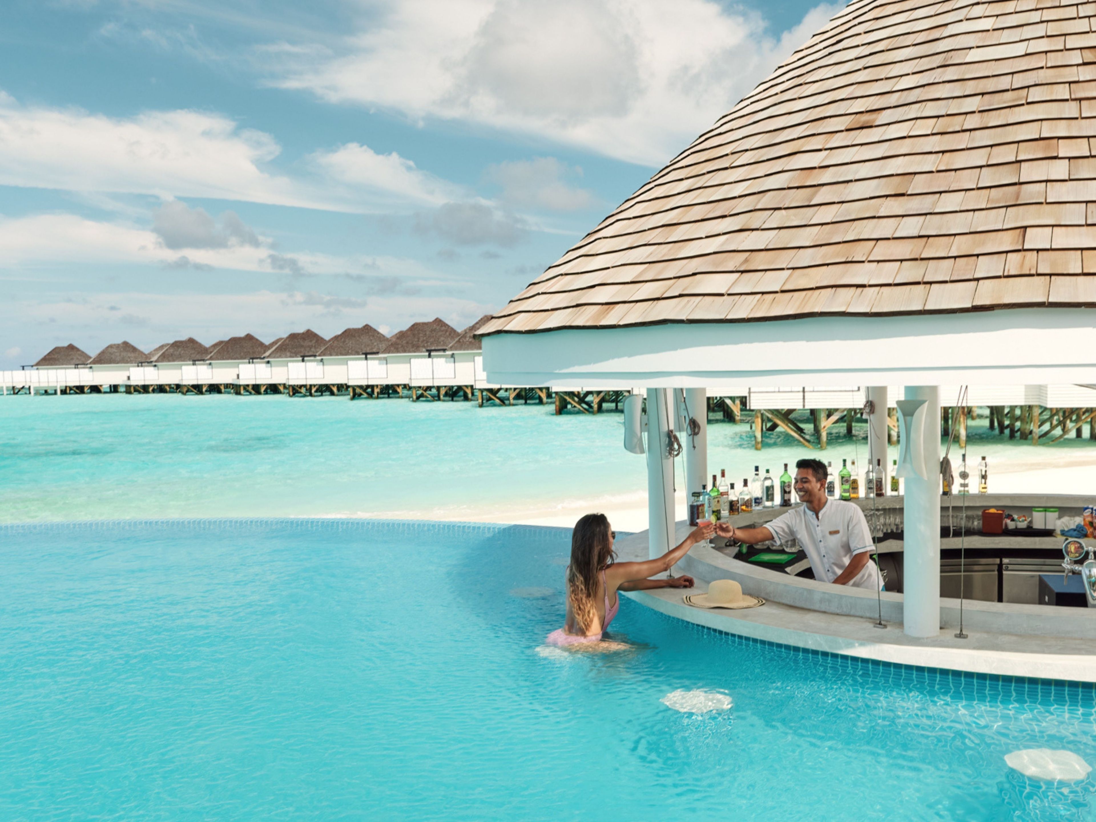 An infinity pool with a couple, a bar, and overwater bungalows in the background.