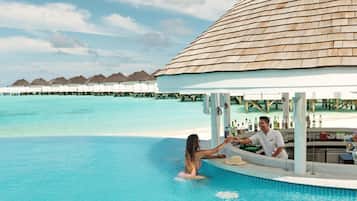 An infinity pool with a couple, a bar, and overwater bungalows in the background.