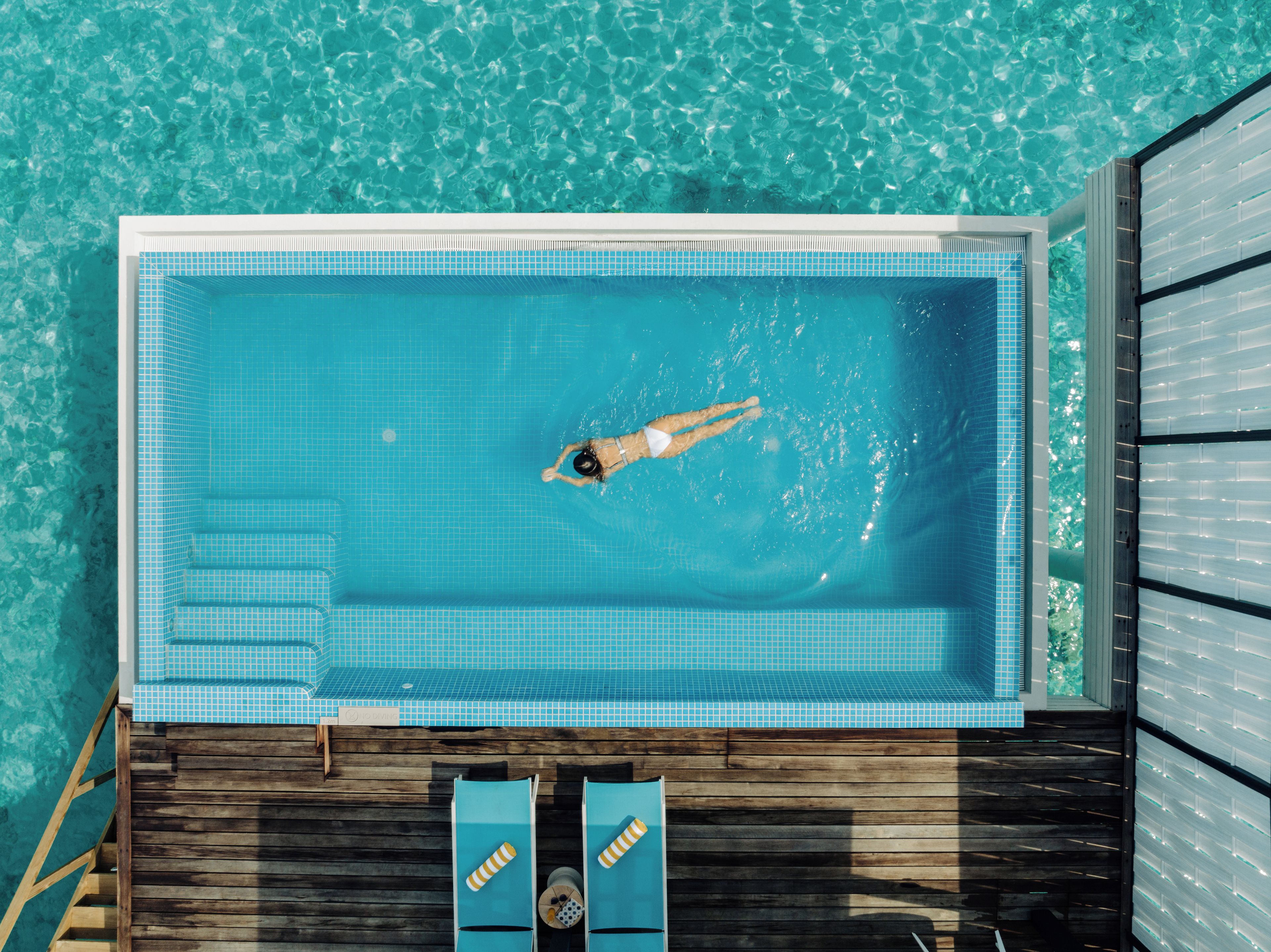 Una piscina rectangular con una zona de solárium con baldosas, un muelle de madera y sillas de descanso.