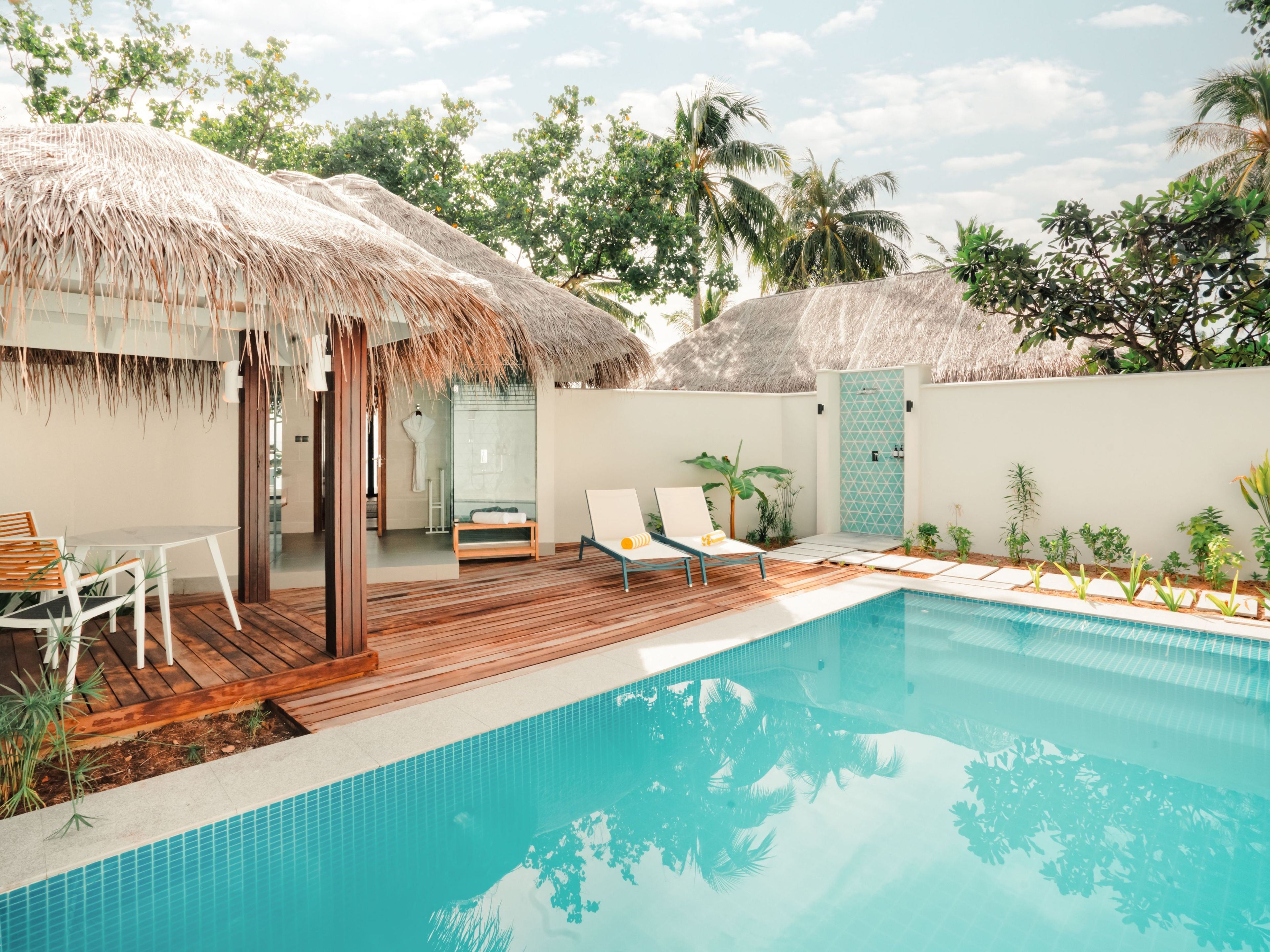 A poolside area with a wooden deck, a glass-enclosed spa, and a thatched-roof structure.