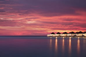 Uma fileira de bangalôs sobre a água, com um mar calmo e céu ao pôr do sol.