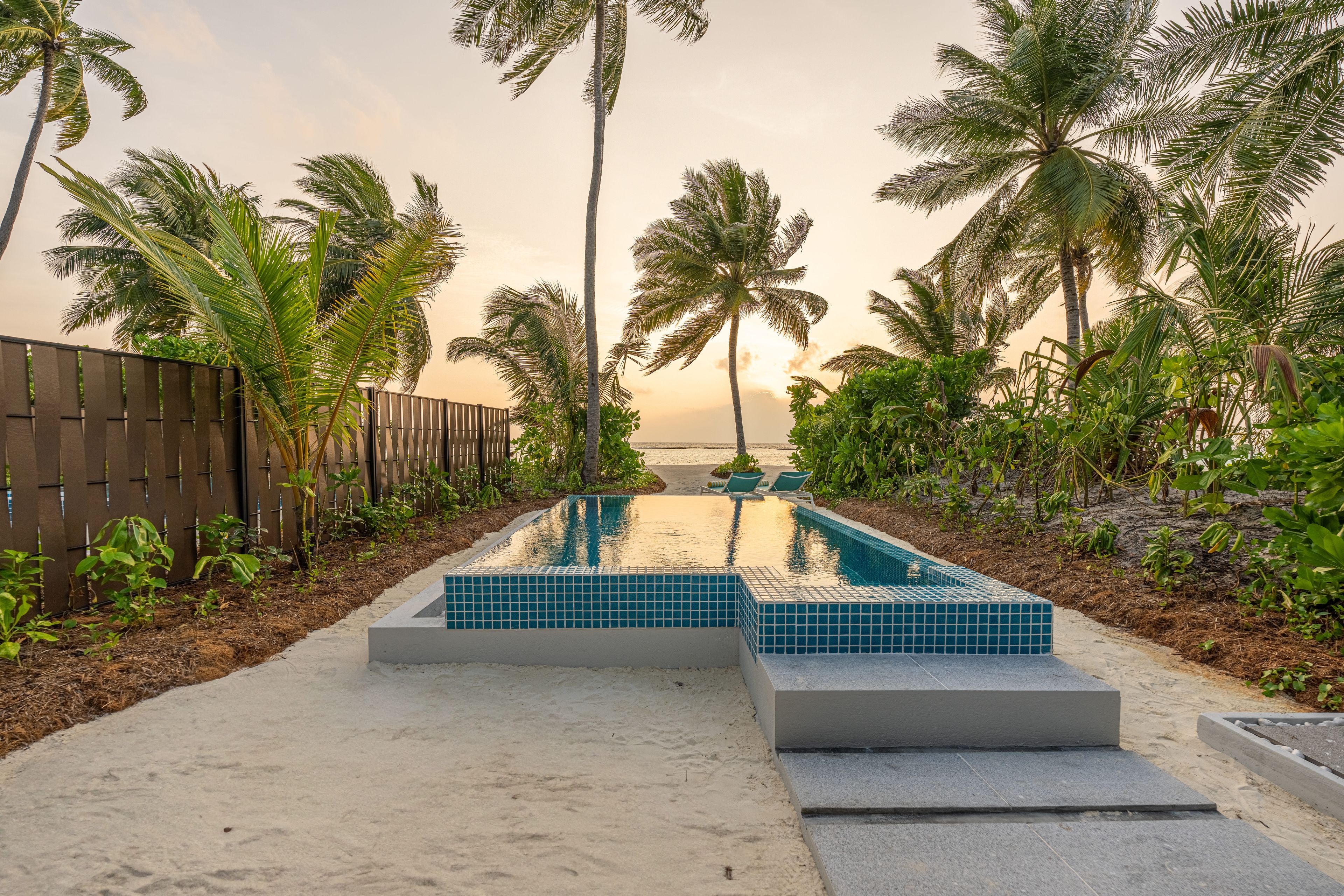 A swimming pool with tiled steps, surrounded by palm trees and a sandy area.