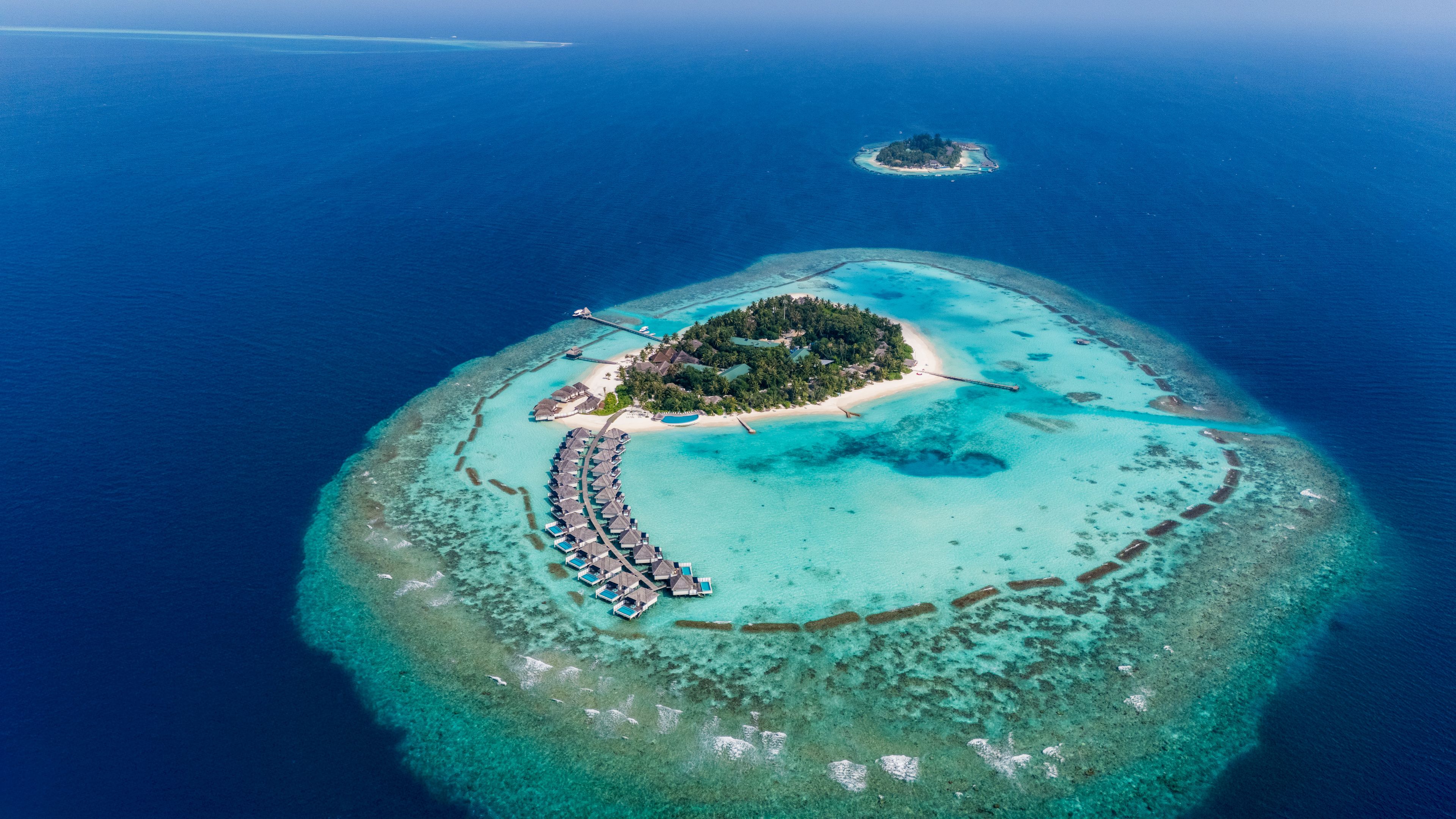 An aerial view of a tropical island resort with a crescent-shaped beach, overwater bungalows, and a small island in the background.