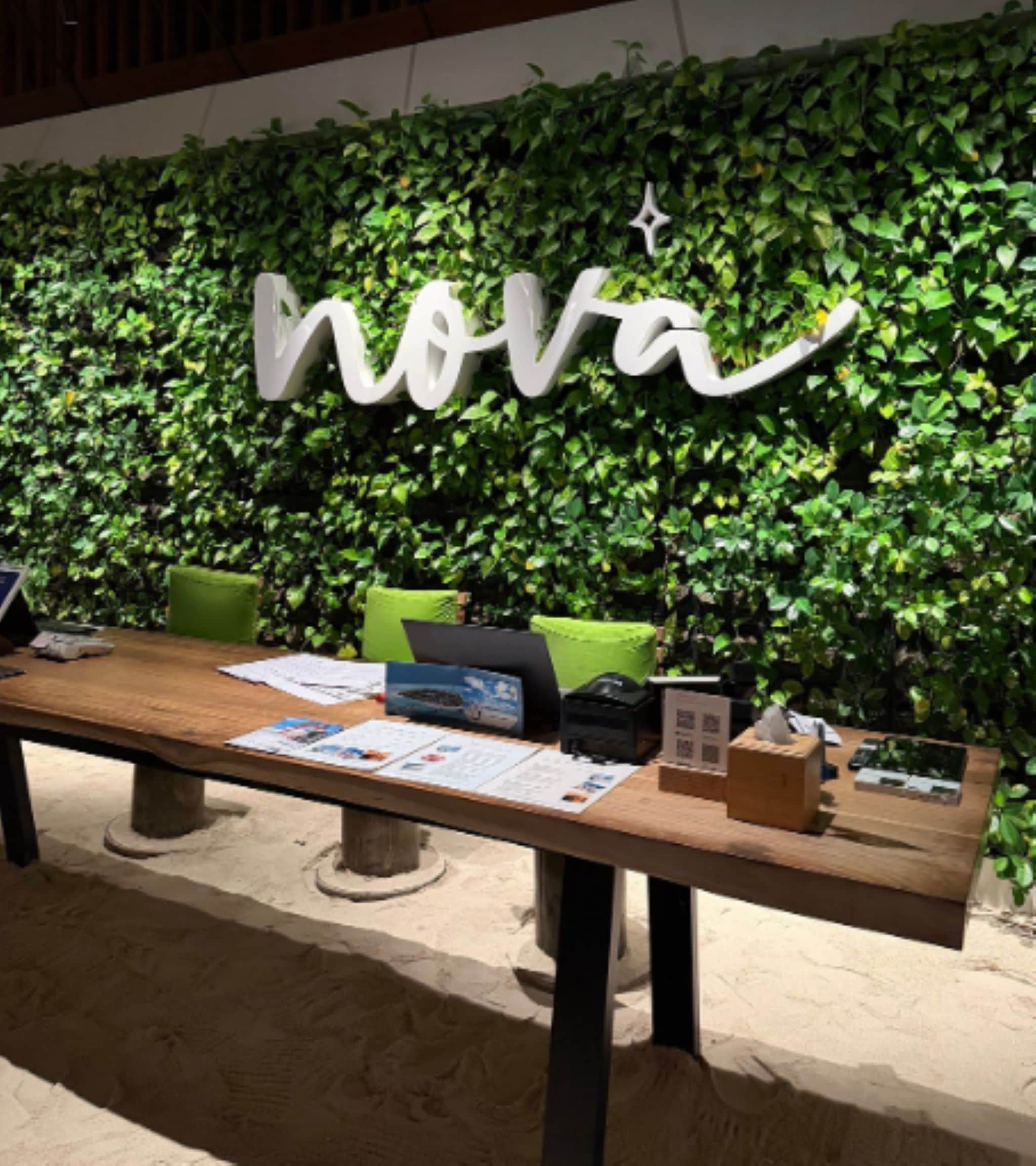 A table with a laptop, papers, and a box, set against a wall with a green leafy backdrop and a white sign with the word 'nova'.
