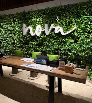A table with a laptop, papers, and a box, set against a wall with a green leafy backdrop and a white sign with the word 'nova'. - Nova Maldives (Malé)