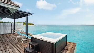 A wooden deck with a hot tub, lounge chairs, and a covered seating area overlooking a clear blue sea.