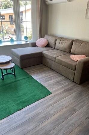 Een woonkamer met een hoekbank, een ronde salontafel, een groen vloerkleed en een raam met uitzicht op gebouwen.