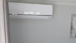 Een kamer met een witte airconditioner aan de muur, een bruine bank met een roze kussen en een ingelijst schilderij.