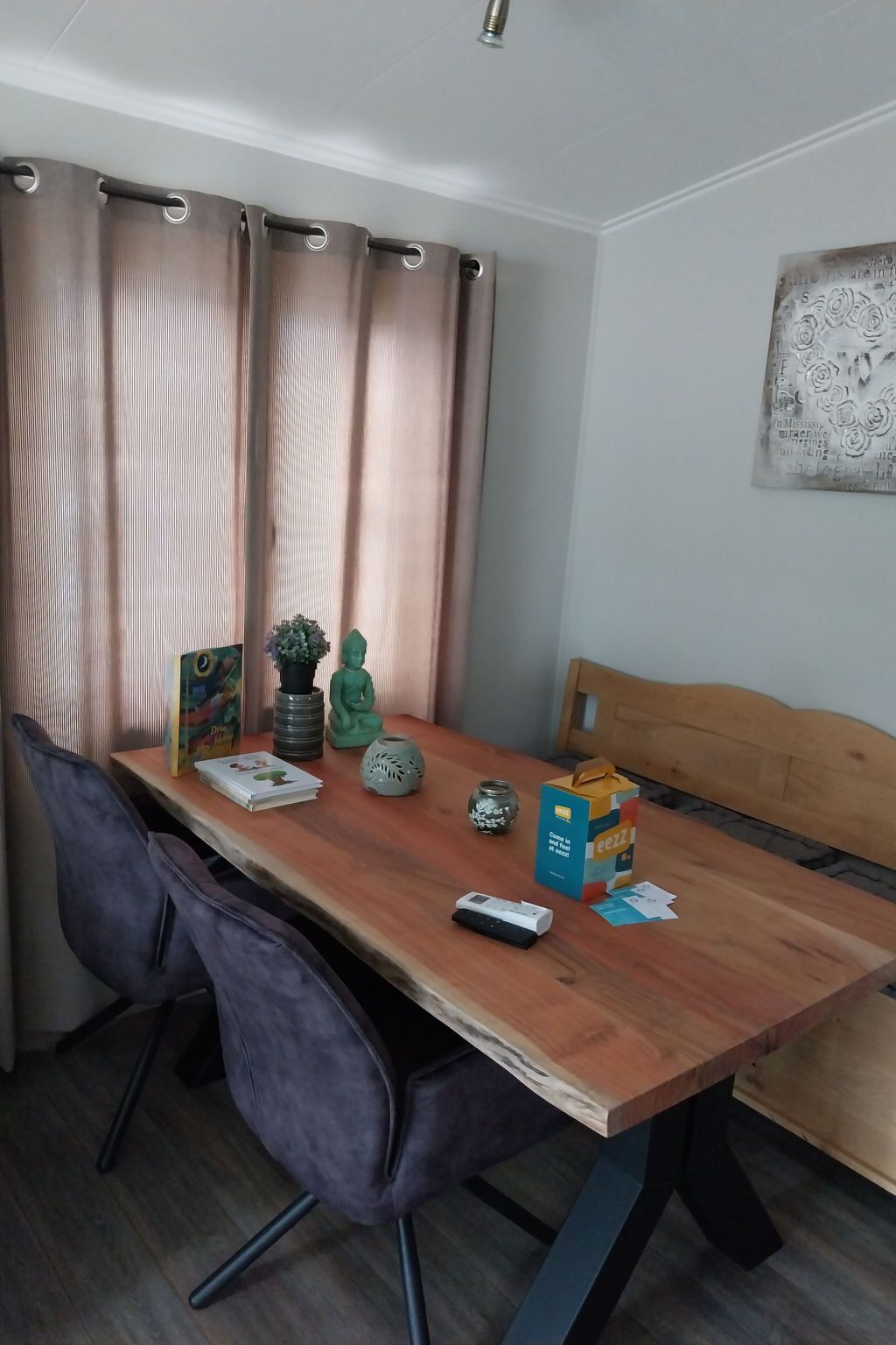 Een eethoek met een houten tafel, twee stoelen, een bankje, een kleine potplant, een decoratieve vaas, een boek, een afstandsbediening en een vruchtensapje.