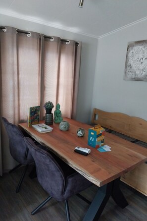 Een eethoek met een houten tafel, twee stoelen, een bankje, een kleine potplant, een decoratieve vaas, een boek, een afstandsbediening en een vruchtensapje.
