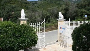 Ein verschlossenes Tor mit weißem Zaun und zwei Löwenstatuen, umgeben von Grünpflanzen und Hügeln.