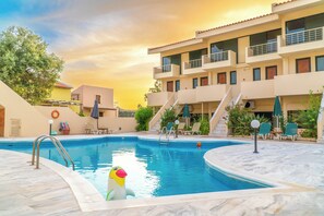 A swimming pool with a lifeguard stand, lounge chairs, and a building with balconies. - Orestis (Chania)