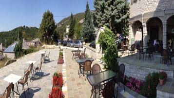 An outdoor patio with stone flooring, tables, and chairs, surrounded by greenery and a stone building with arches.