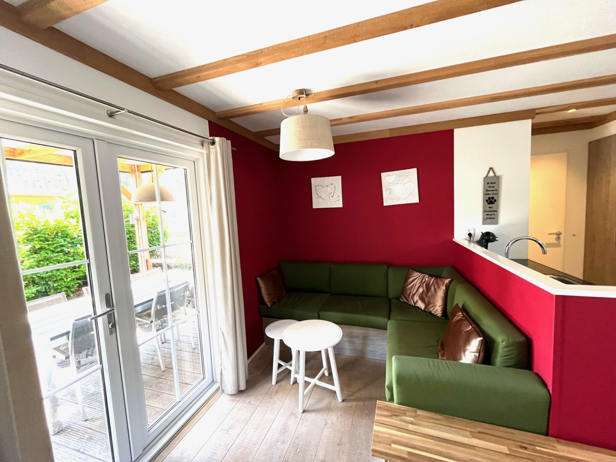 Een kamer met een groene bank, een witte kruk, een houten tafel, een raam met gordijnen en een rode muur met twee ingelijste foto's.