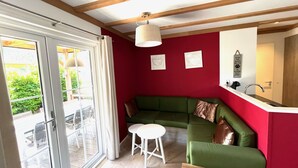 Een kamer met een groene bank, een witte kruk, een houten tafel, een raam met gordijnen en een rode muur met twee ingelijste foto's.