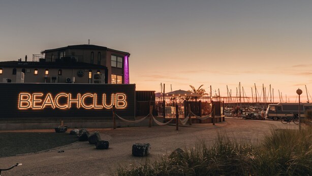 Ein Strandclub mit leuchtendem Neonzeichen, ein Gebäude mit Balkon und ein Yachthafen im Hintergrund.