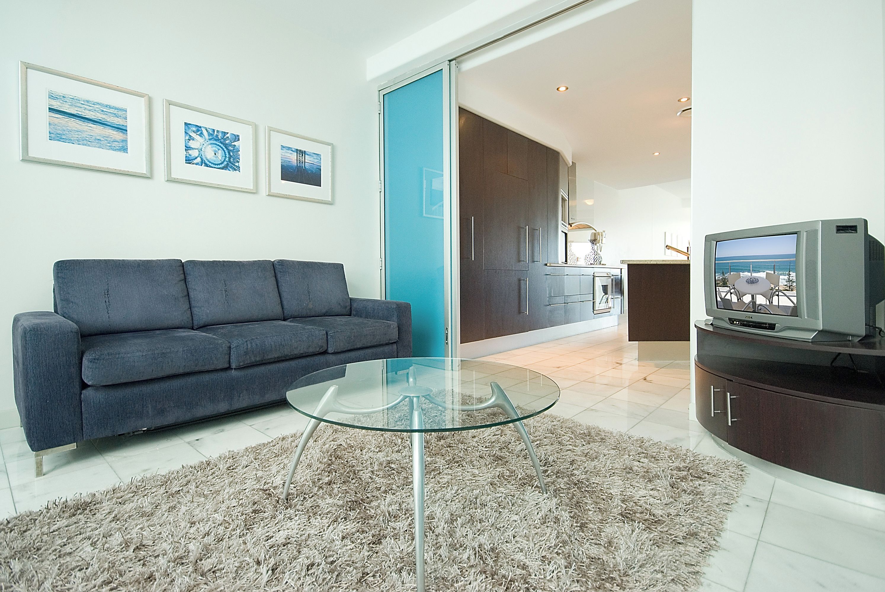A modern living room with a grey sofa, a glass coffee table, and a TV on a wooden cabinet. There are framed pictures on the wall and a kitchen area is visible through an open door.