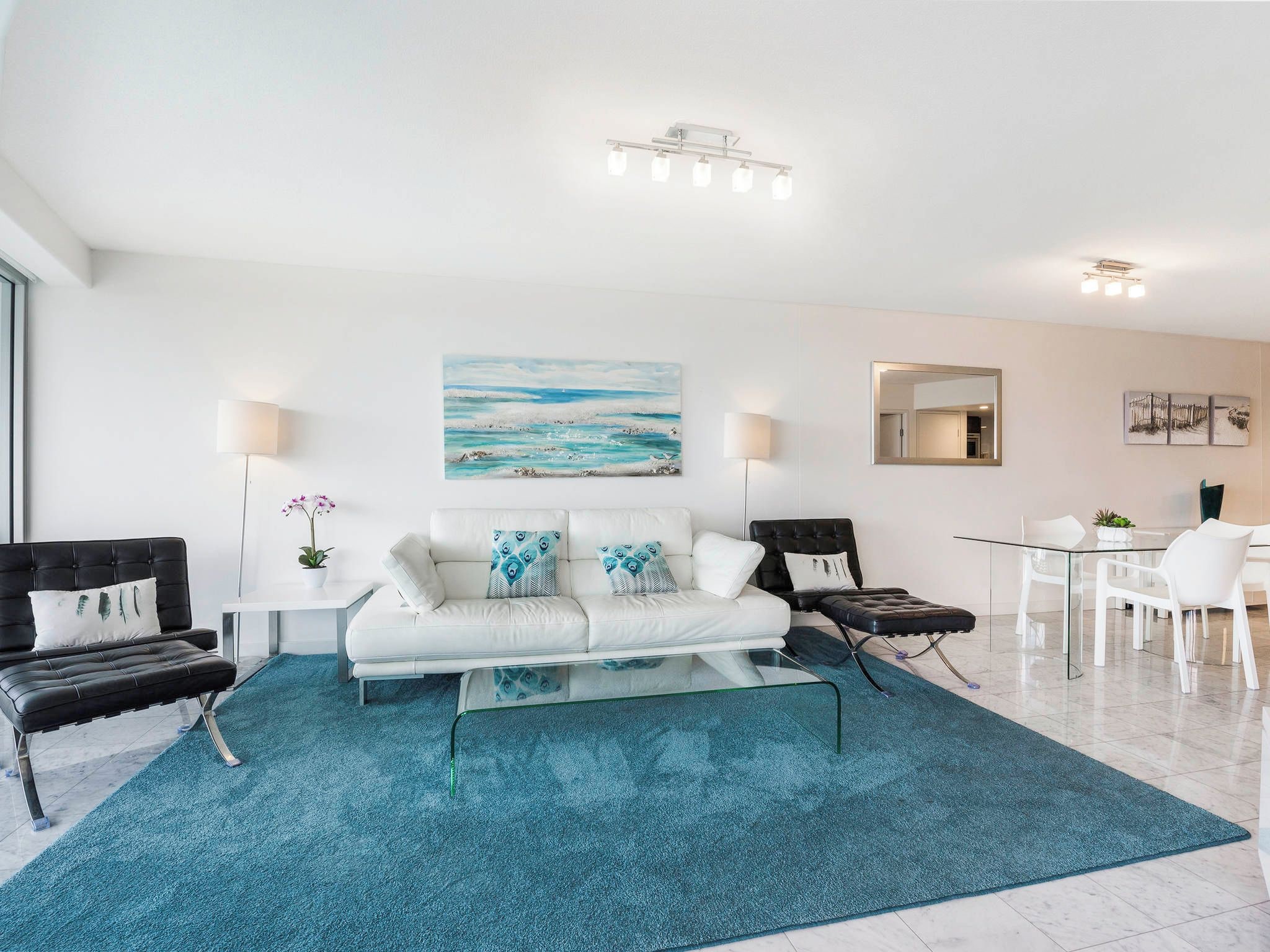 A modern living room with a white sofa, a glass coffee table, and a blue rug. There is a dining area with a glass table and white chairs.