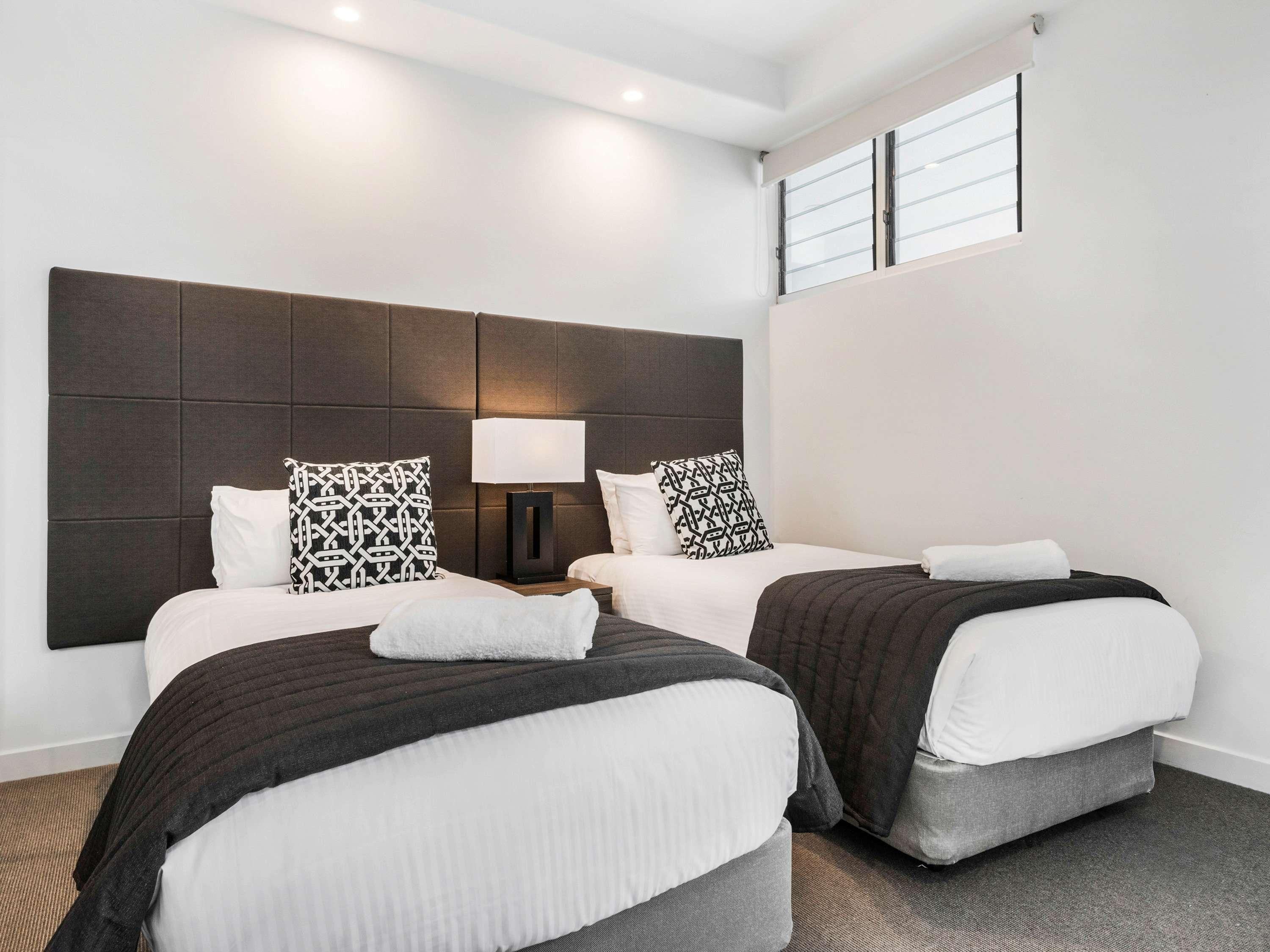 Une chambre d’hôtel avec deux lits, une grande tête de lit, une table de chevet avec une lampe et une fenêtre avec des stores.