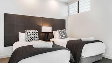 Une chambre d’hôtel avec deux lits, une grande tête de lit, une table de chevet avec une lampe et une fenêtre avec des stores.
