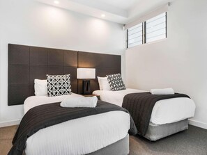 Hotellihuone, jossa on kaksi sänkyä, suuri päätyseinä, yöpöytä lampulla ja ikkuna, jossa on sälekaihtimet.