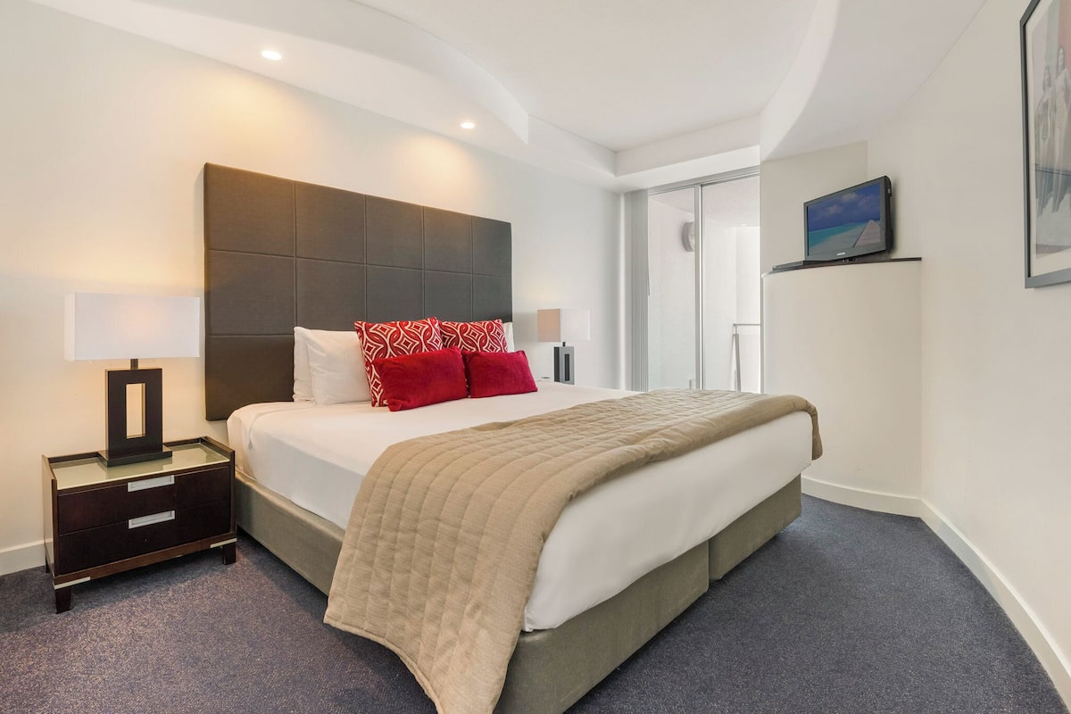 Una habitación de hotel moderna con una cama grande, una mesita de noche con lámpara, un armario y un televisor montado en la pared.