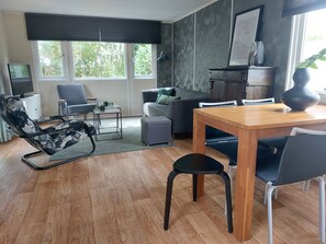 Ein modernes Wohnzimmer mit einer Couch, einem Esstisch, einem Sessel, einem Fernseher und einem Fenster mit Blick auf Bäume.