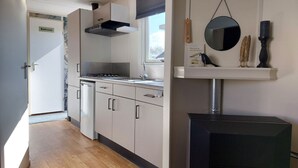 Een moderne keuken met een fornuis, spoelbak en kastjes. Er is een raam met zonwering, een spiegel aan de muur en een zwarte verwarming.