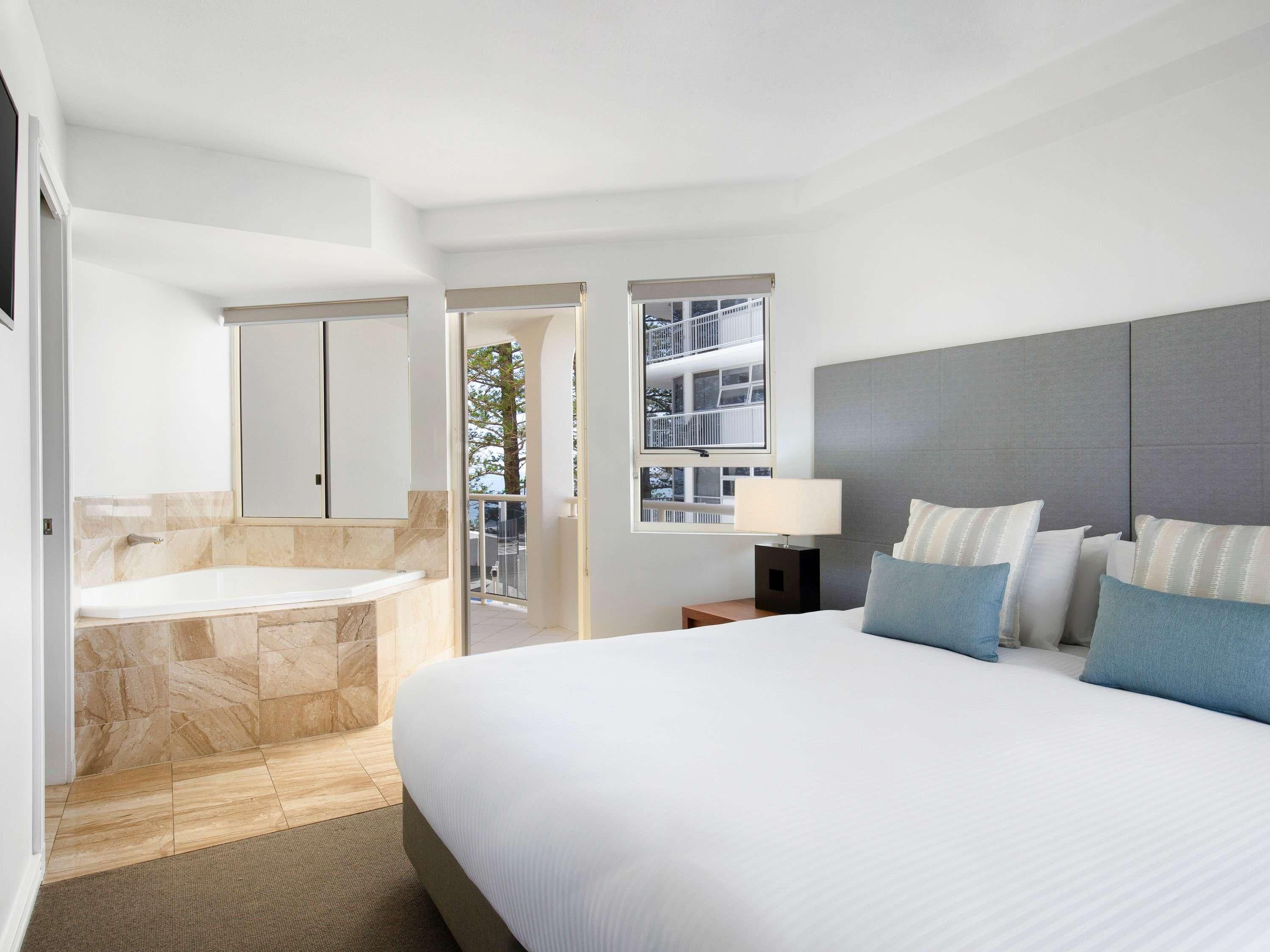 A modern hotel room with a large bed, a separate bathroom with a bathtub, and a balcony with a view of the building.