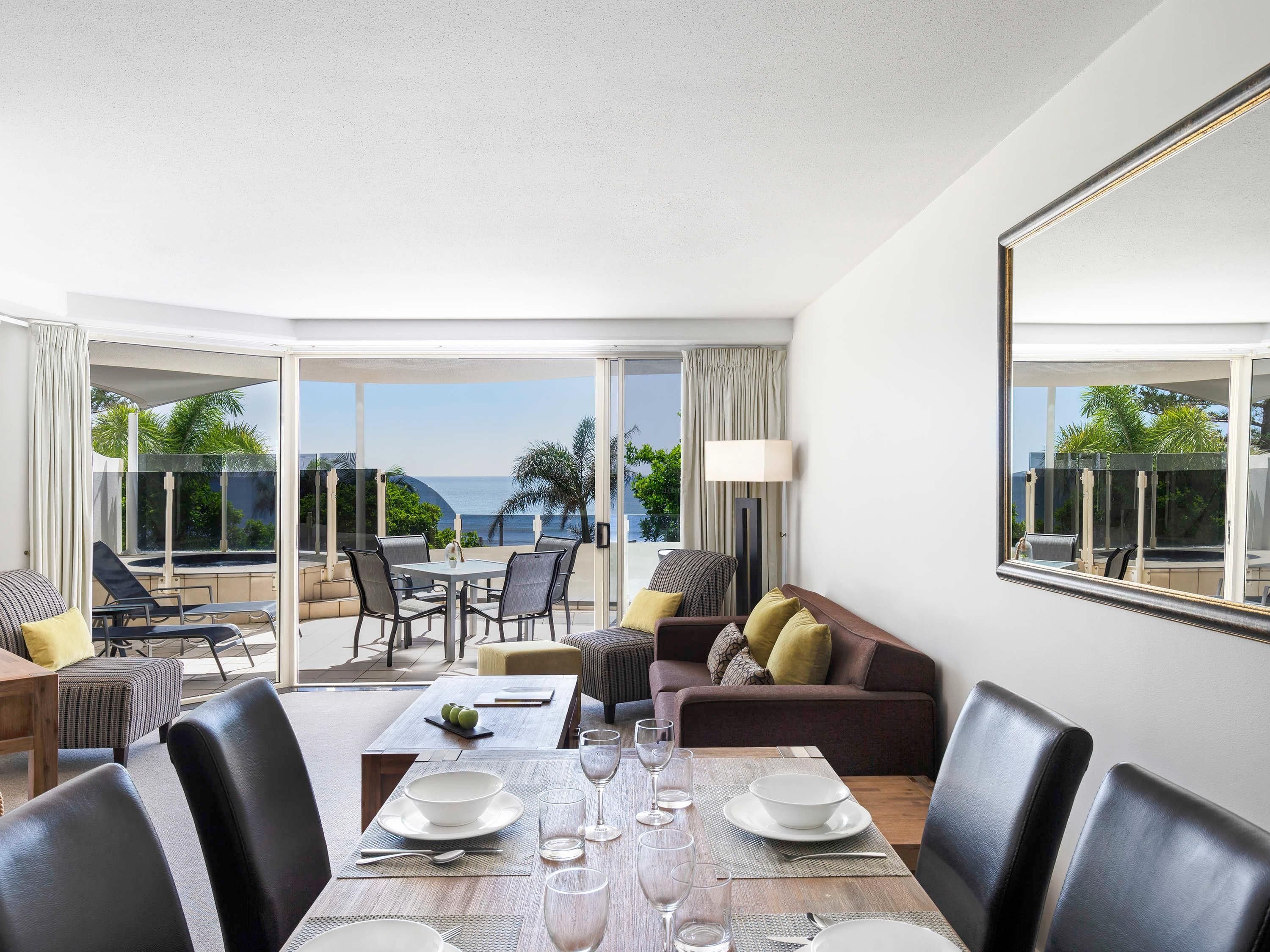 A hotel room with a dining area, a sofa, and a large window with a view of the sea.