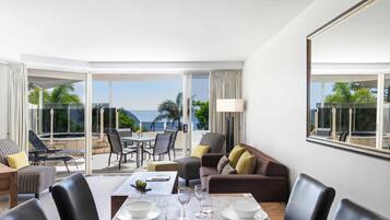 A hotel room with a dining area, a sofa, and a large window with a view of the sea.